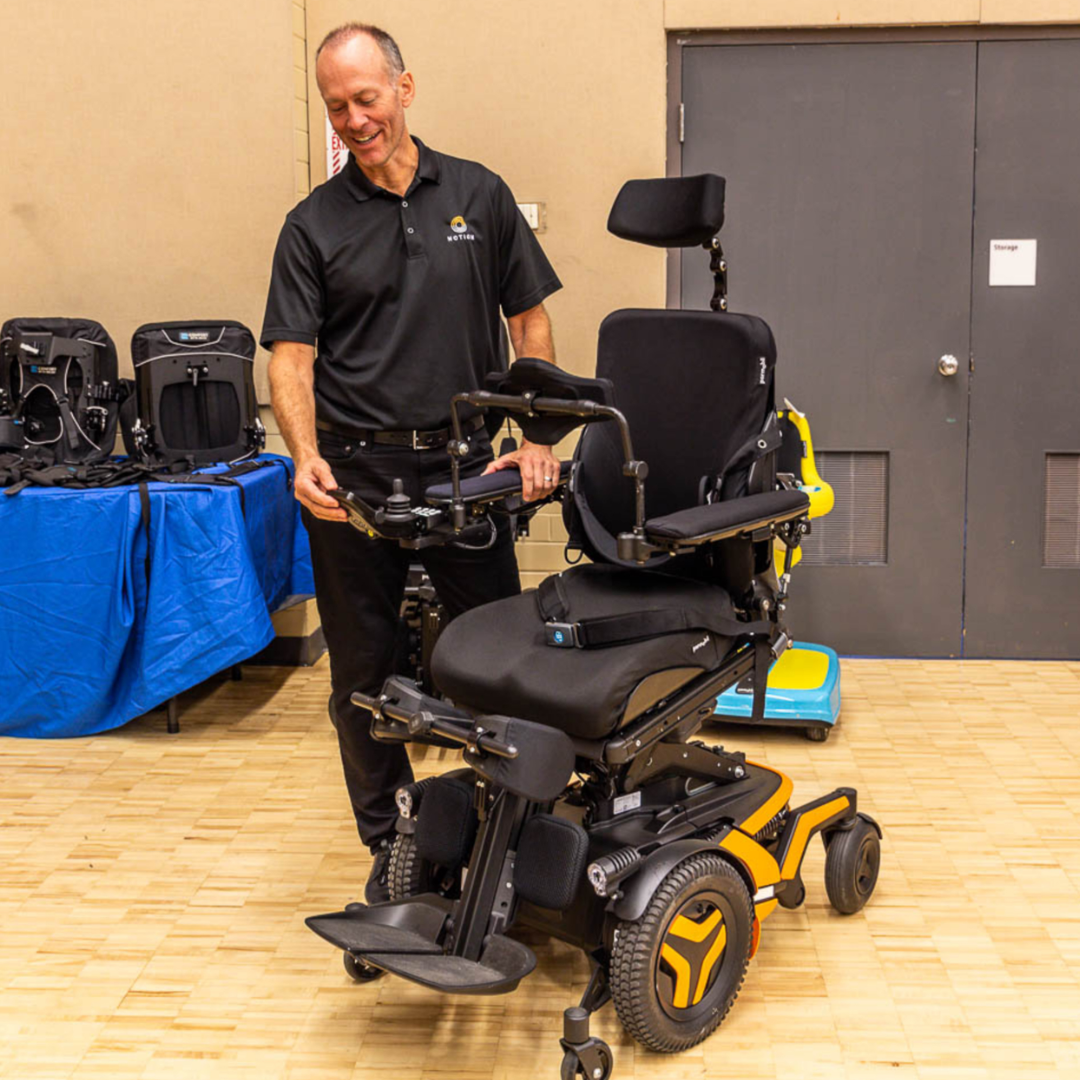 Motion Employee standing next to a power wheelchair with a yellow base. The employee is looking downwards with his hand on the joystick